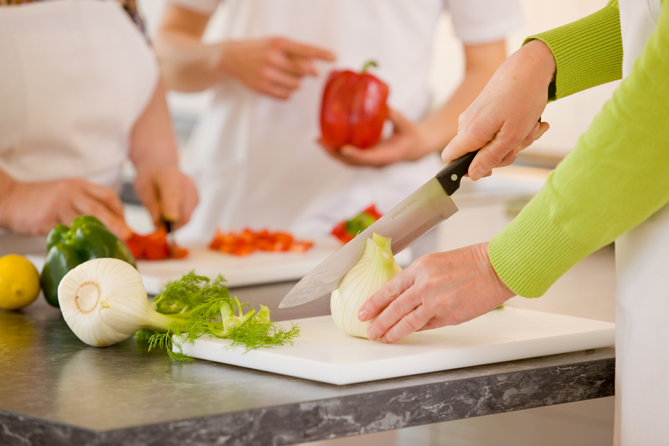 Hände von drei Personen, die an einer Arbeitsplatte auf Brettchen Fenchel und Paprika schneiden